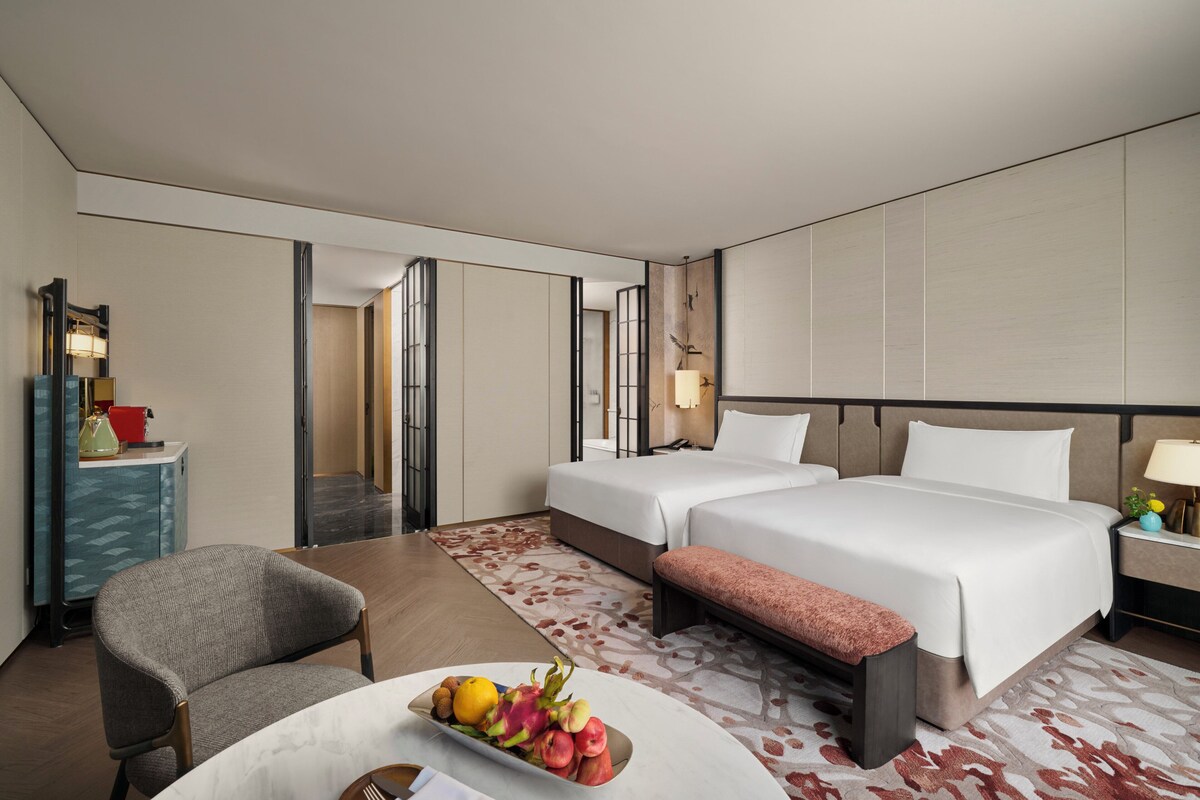 Habitación de hotel con dos camas, un escritorio con silla, un frutero y una mesita de noche con lámpara.