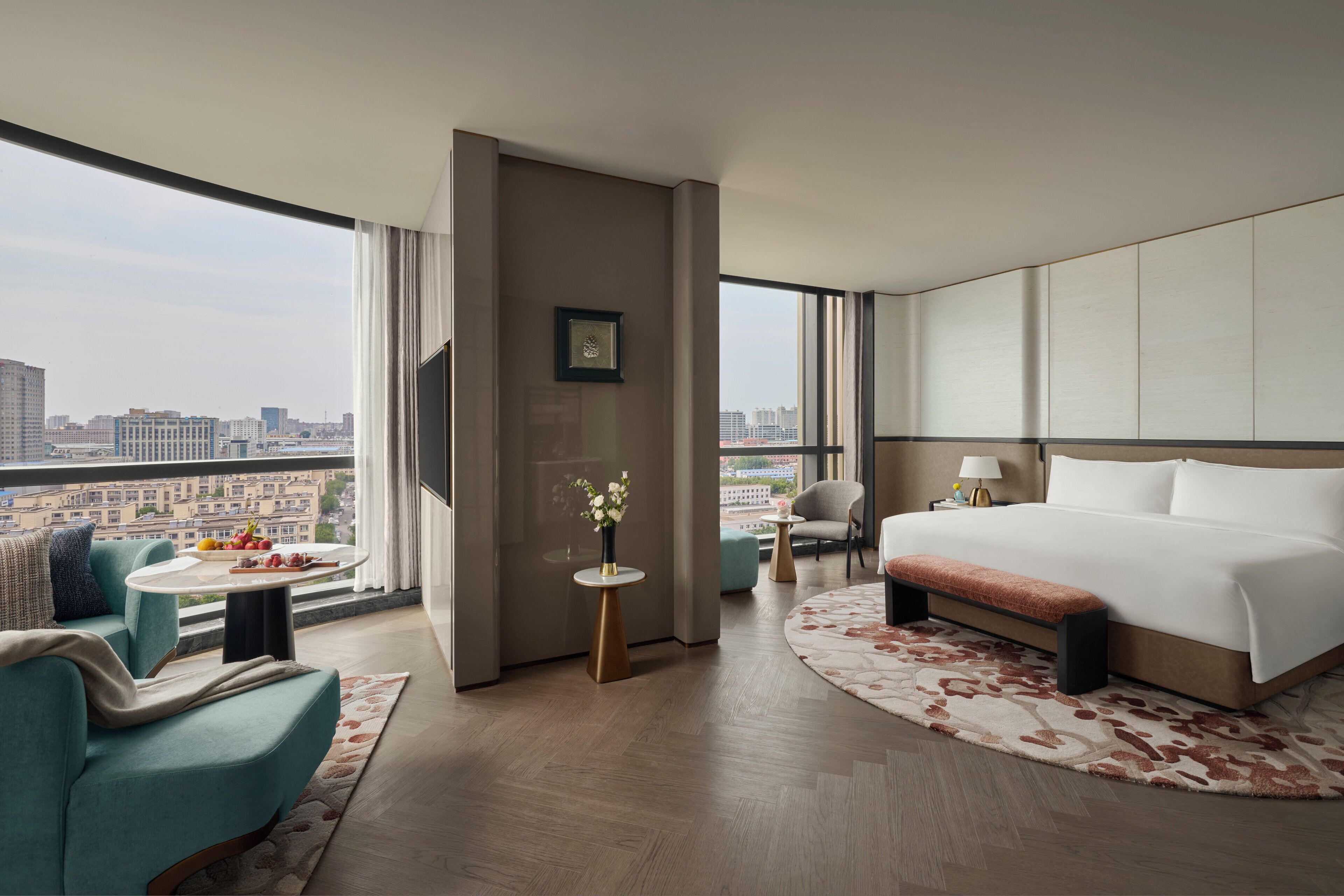 A hotel room with a large bed, a sofa, a small round table with a vase of flowers, and a view of the cityscape.
