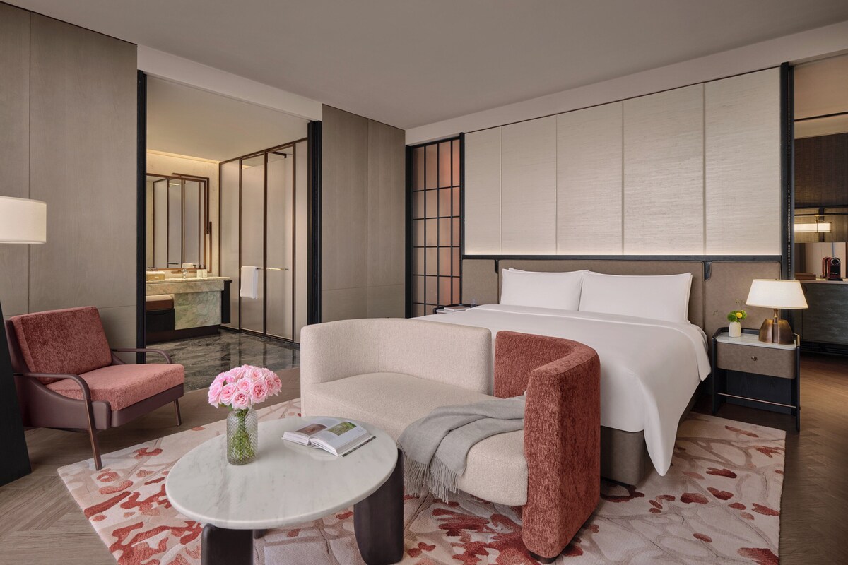 Habitación de hotel con una cama grande, un área de estar con una mesa y sillas, un baño con espejo y lavamanos, y una mesita de noche con lámpara.