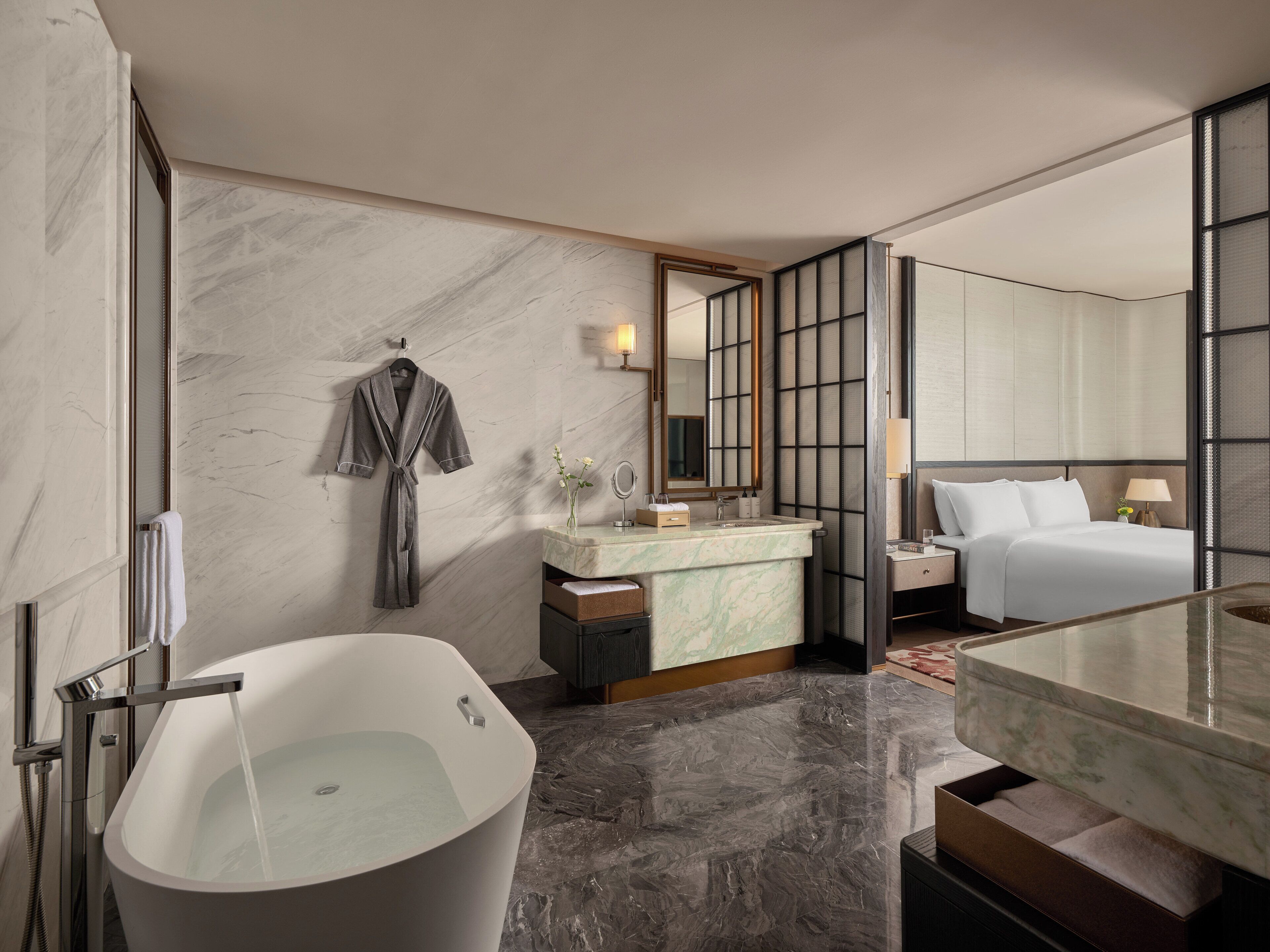 A modern bathroom with a freestanding bathtub, a vanity with a marble countertop, a large mirror, and a bed with white linens.