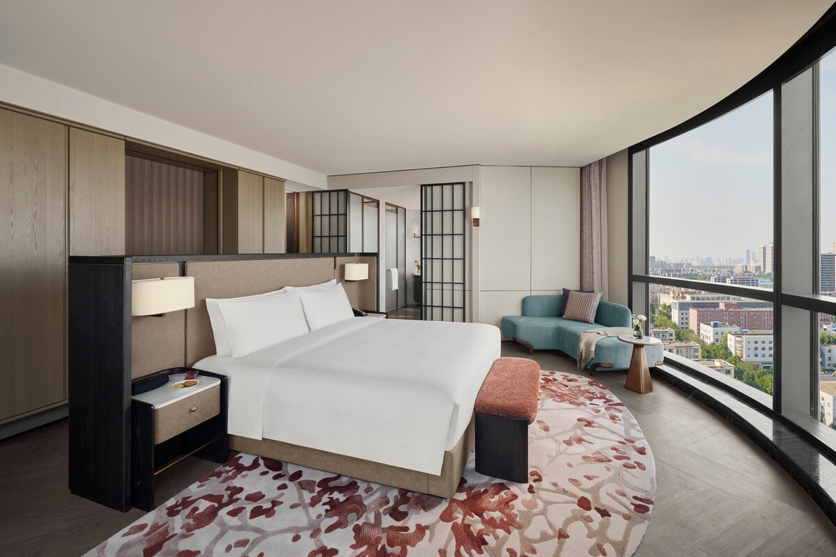 A modern hotel room with a large bed, a seating area with a sofa and armchair, a small side table, and a large window offering a city view.