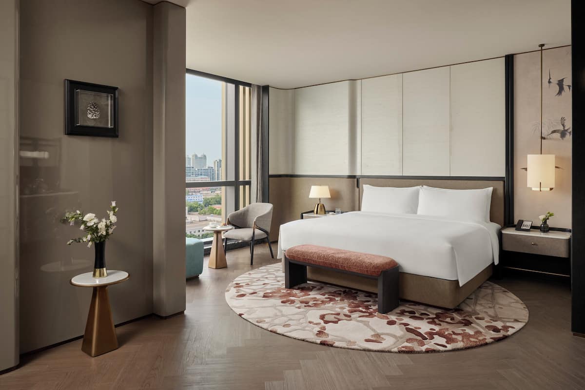 A modern hotel room with a large bed, a round rug, a small table with a vase, a chair, and a view of the city through large windows.