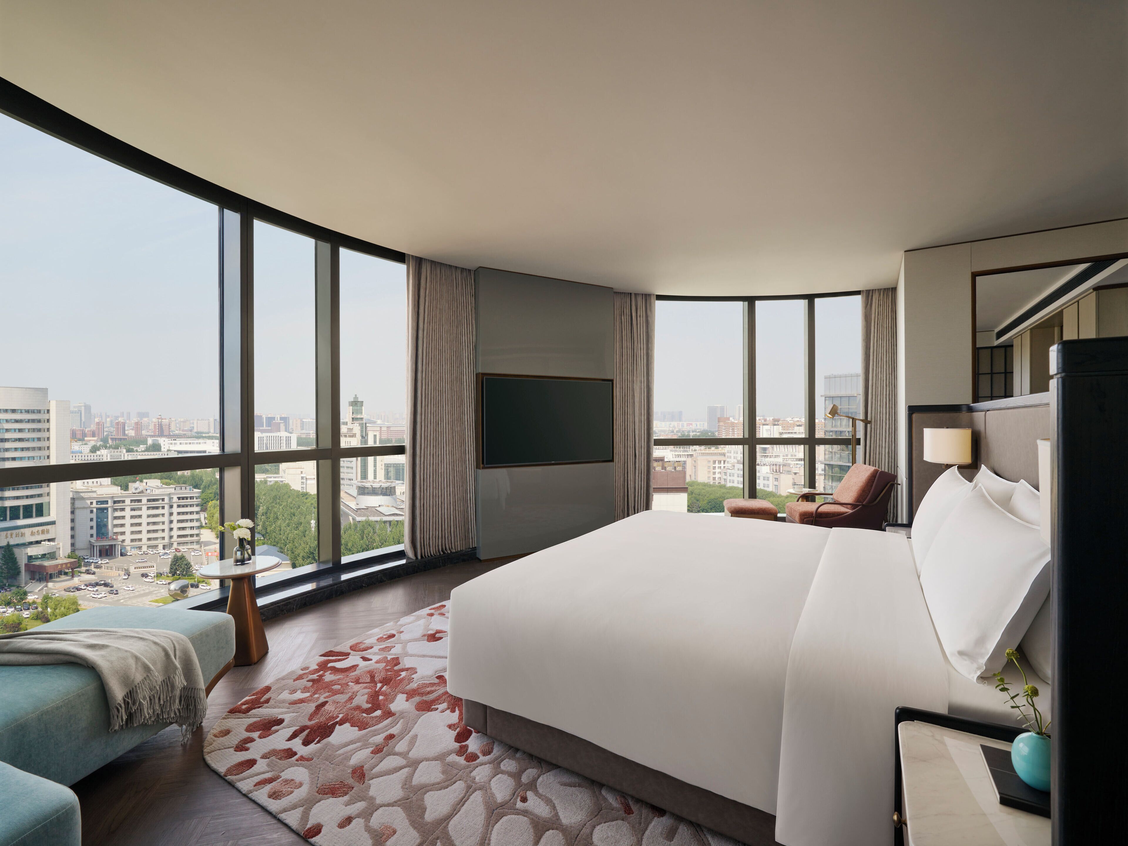 A modern hotel room with a large bed, a seating area with a chair and a small table, a flat-screen TV mounted on the wall, and a city view through large windows.