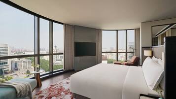 A modern hotel room with a large bed, a seating area with a chair and a small table, a flat-screen TV mounted on the wall, and a city view through large windows.