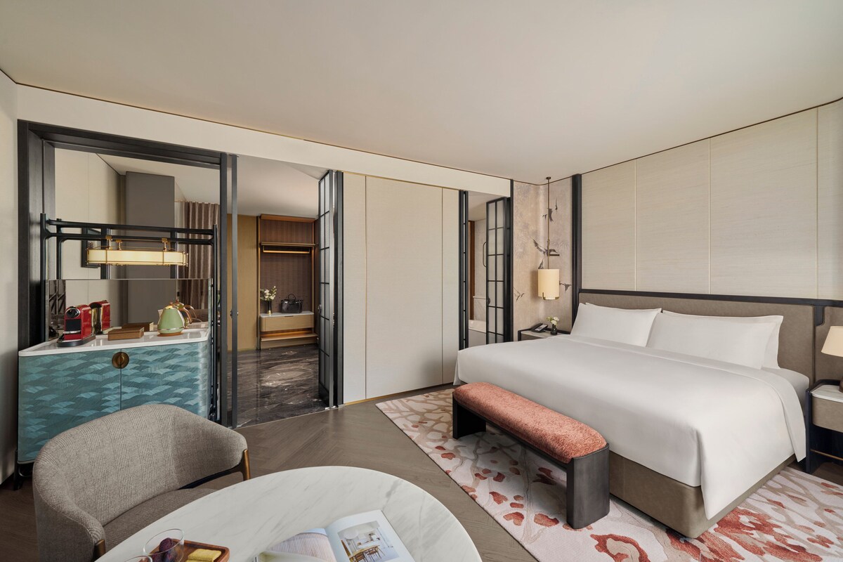 A hotel room with a large bed, a bedside table, a chair, a desk with a lamp, a wardrobe, and a mirror.