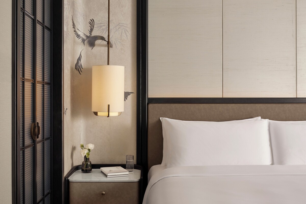 Una habitación de hotel con una cama, una mesita de noche con una lámpara, un jarrón con flores y una pared decorada con un mural de aves.