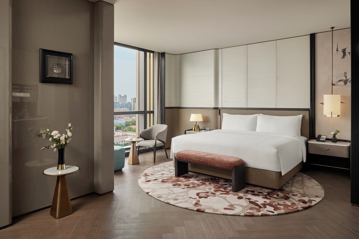 Una habitación de hotel moderna con una cama grande, un tapete redondo, una mesita con un florero, una silla y vistas a la ciudad a través de grandes ventanales.