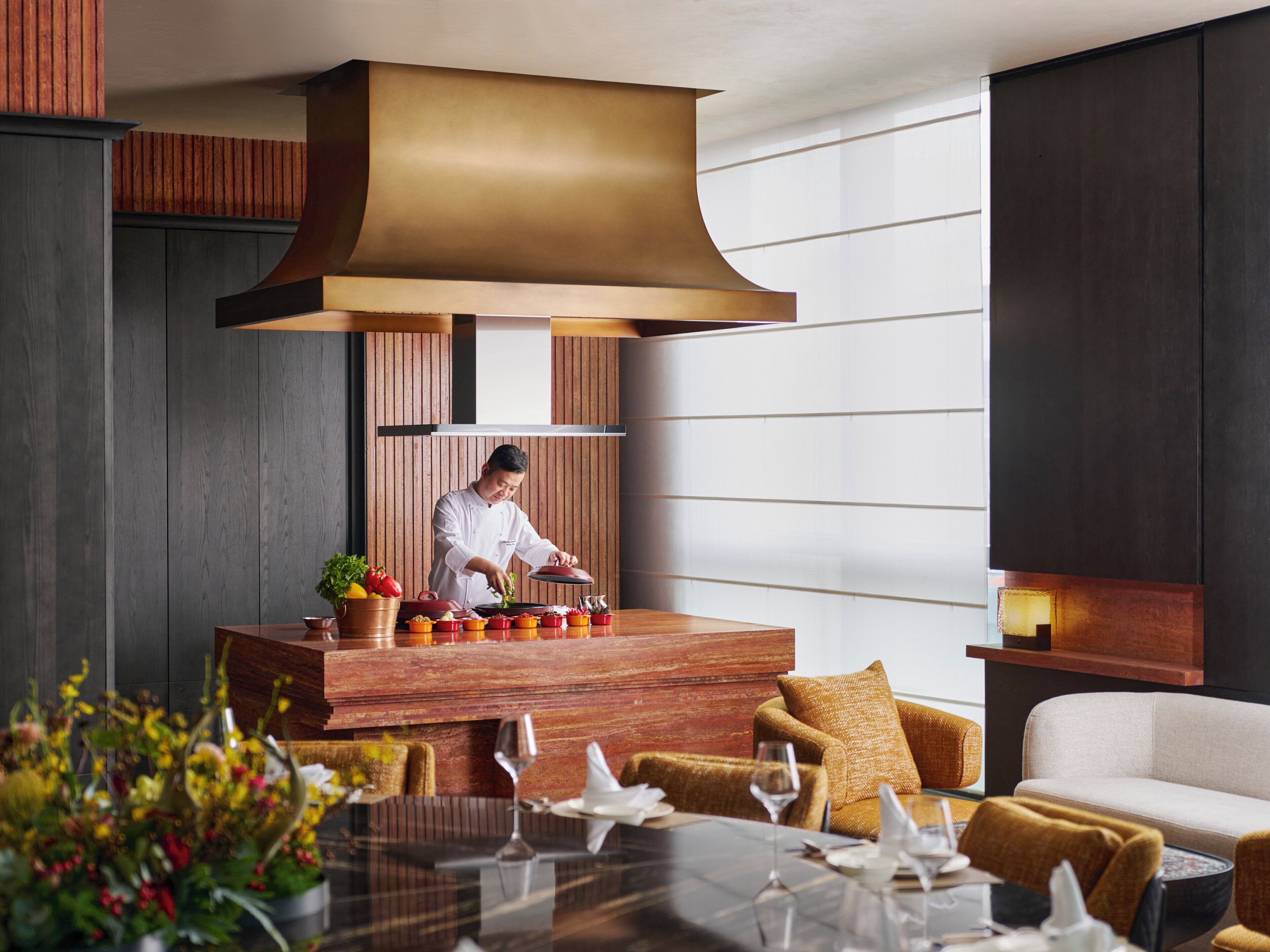 Un espace de restauration moderne avec un chef qui prépare les plats, un bar en bois et un coin salon avec un canapé et des fauteuils.