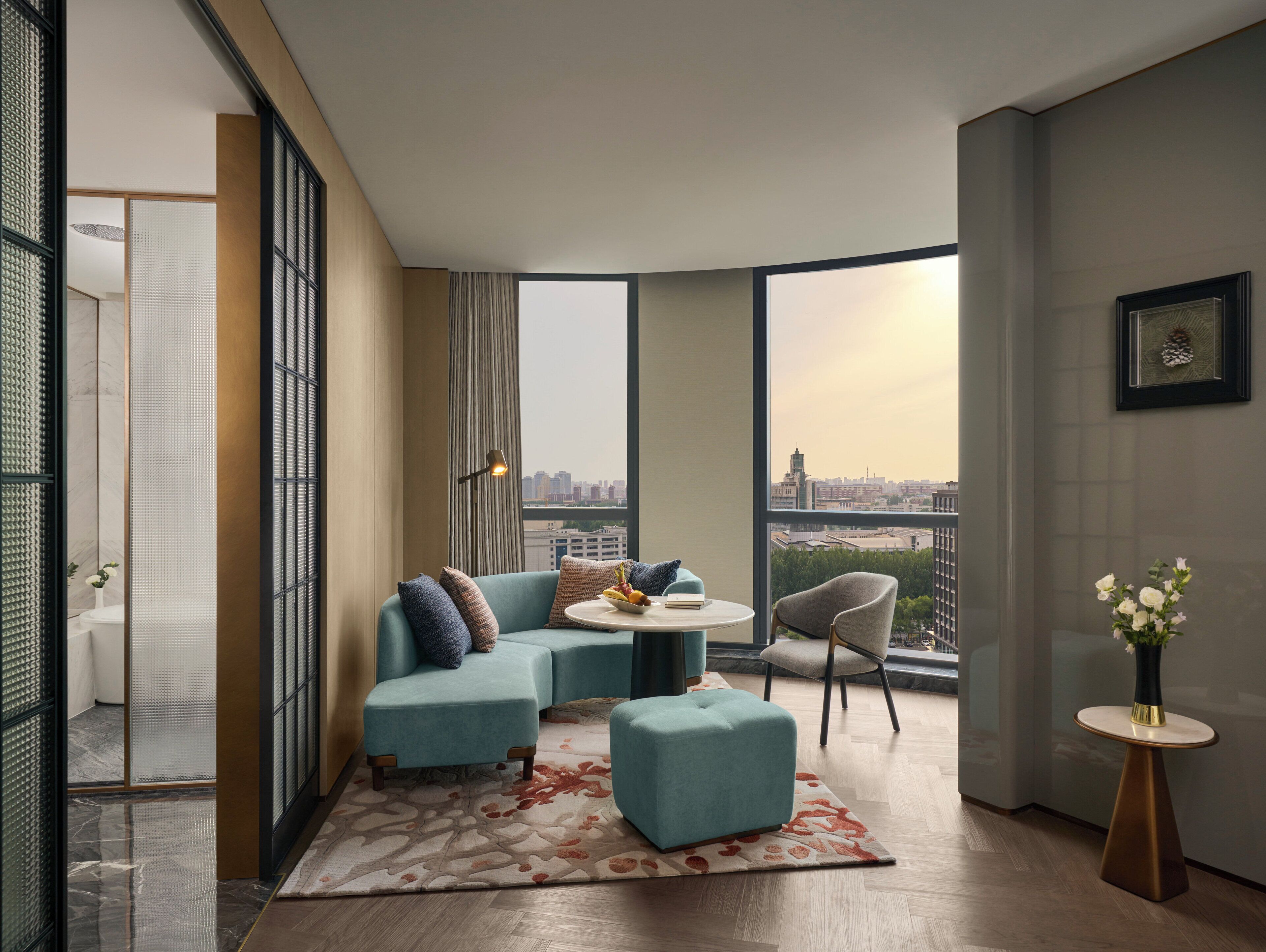 En moderne stue med sofa, skammel og stole, et lille bord med en skål frugt, og udsigt over bybilledet gennem store vinduer.