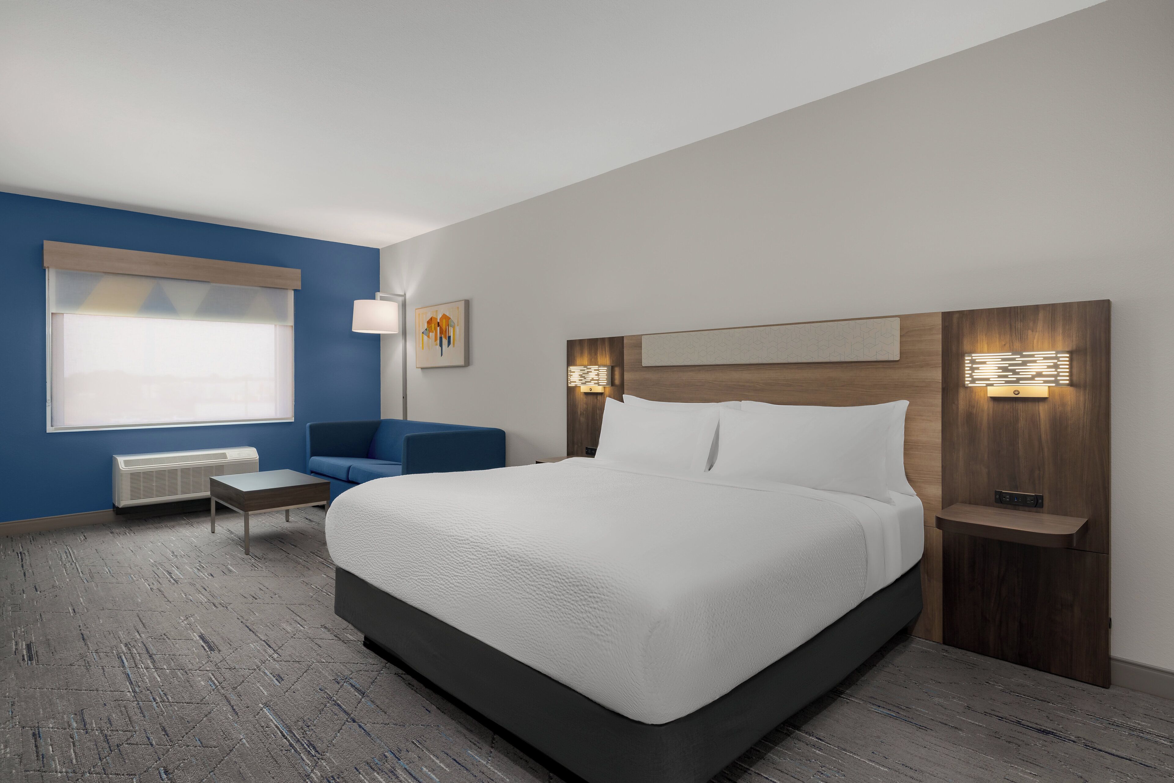 Una habitación de hotel moderna con una cama grande, un muro azul decorativo, una pequeña zona de estar y una ventana con persianas.