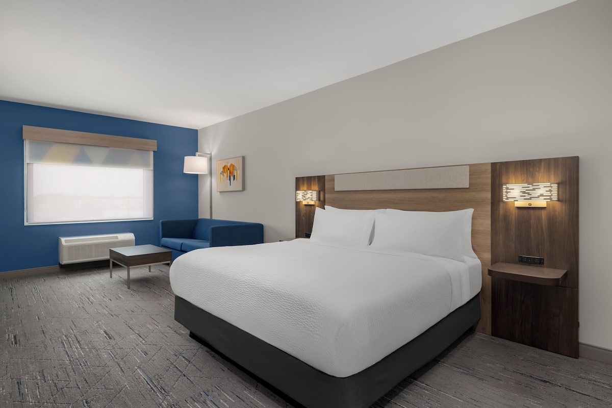 A modern hotel room with a large bed, a blue accent wall, a small seating area, and a window with blinds.