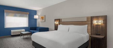 A modern hotel room with a large bed, a seating area with a sofa and armchair, a small table, and a window with blinds.