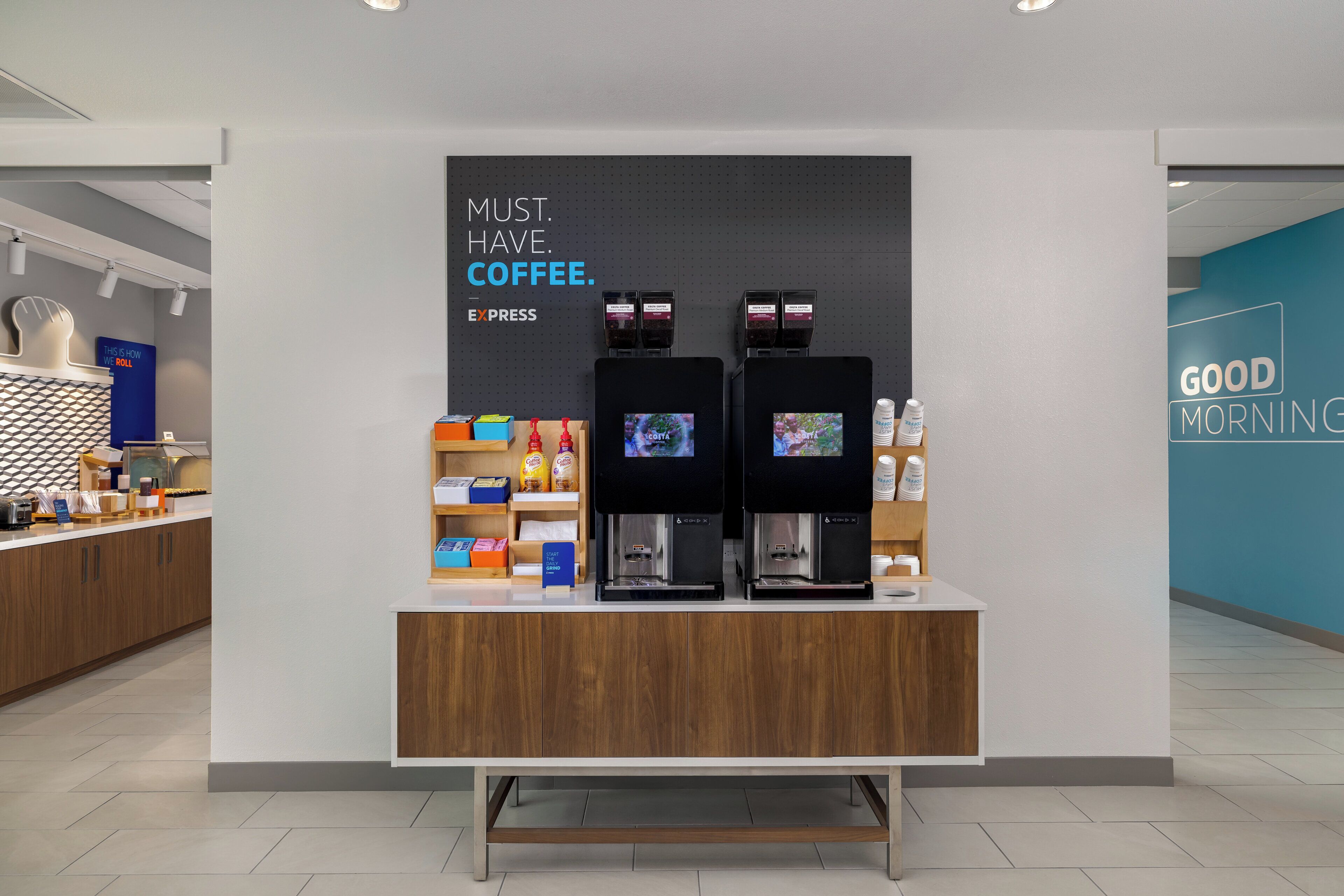 A coffee station with two espresso machines, a sign that says 'MUST. HAVE. COFFEE. EXPRESS,' and a 'GOOD MORNING' sign on the wall.