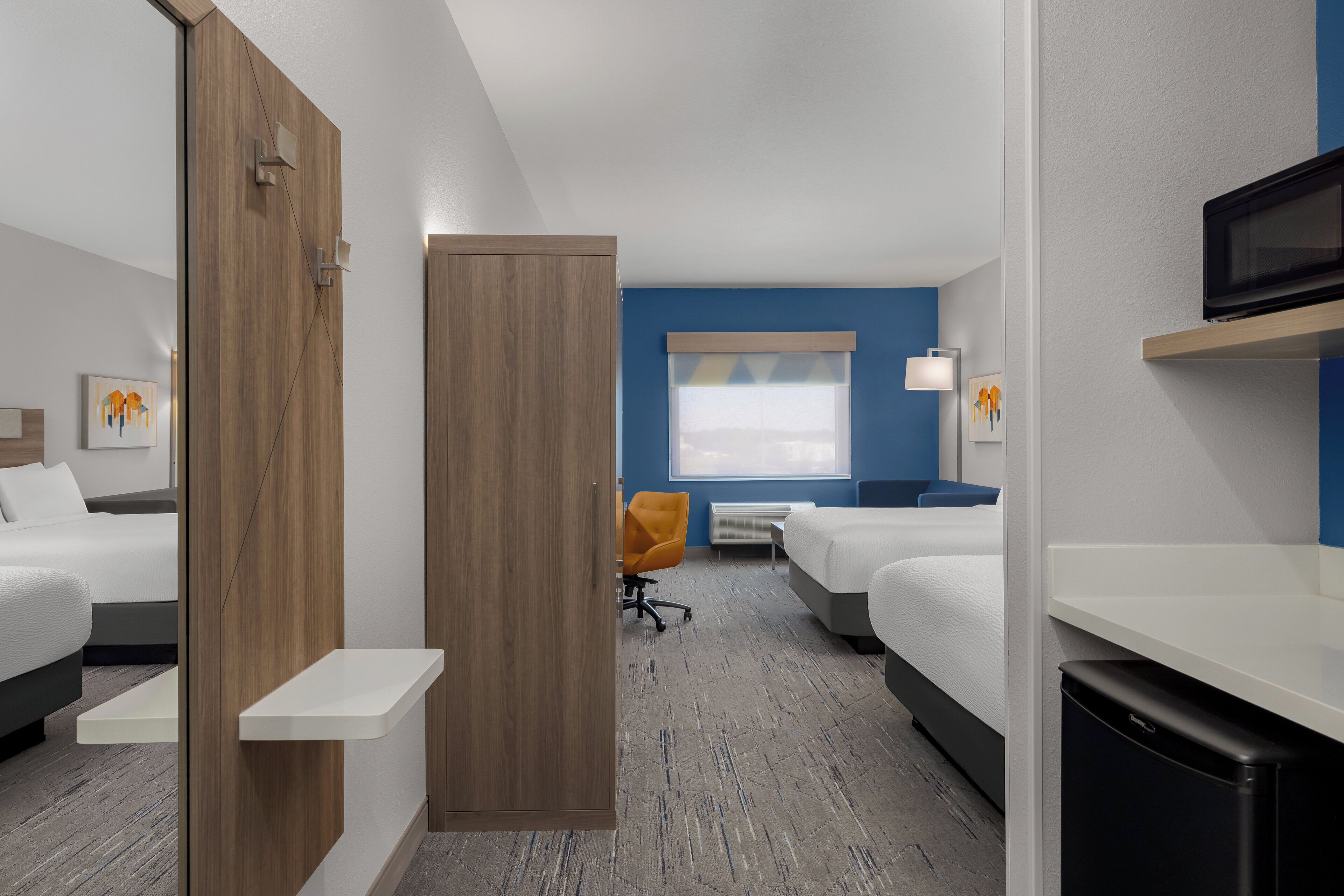 A hotel room with two beds, a desk, a chair, a microwave, and a window with blinds.