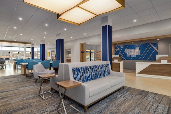 A modern hotel lobby with a reception desk, seating area, and a food kiosk.