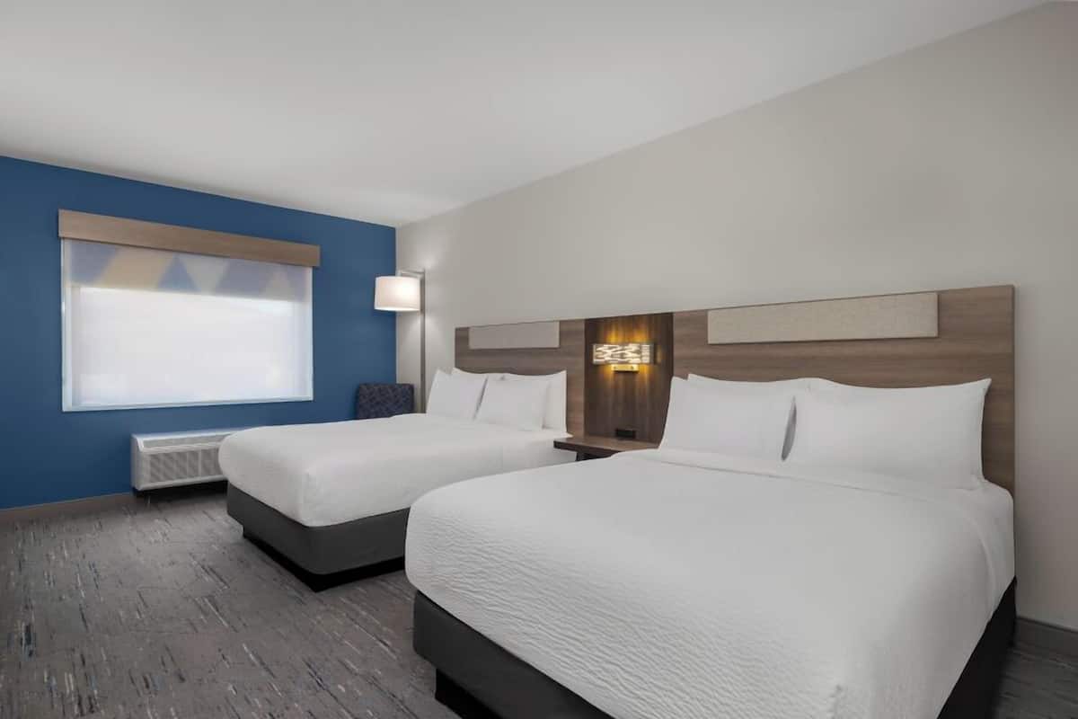 A hotel room with two beds, a large window with blinds, a standing lamp, and a wall-mounted light fixture.