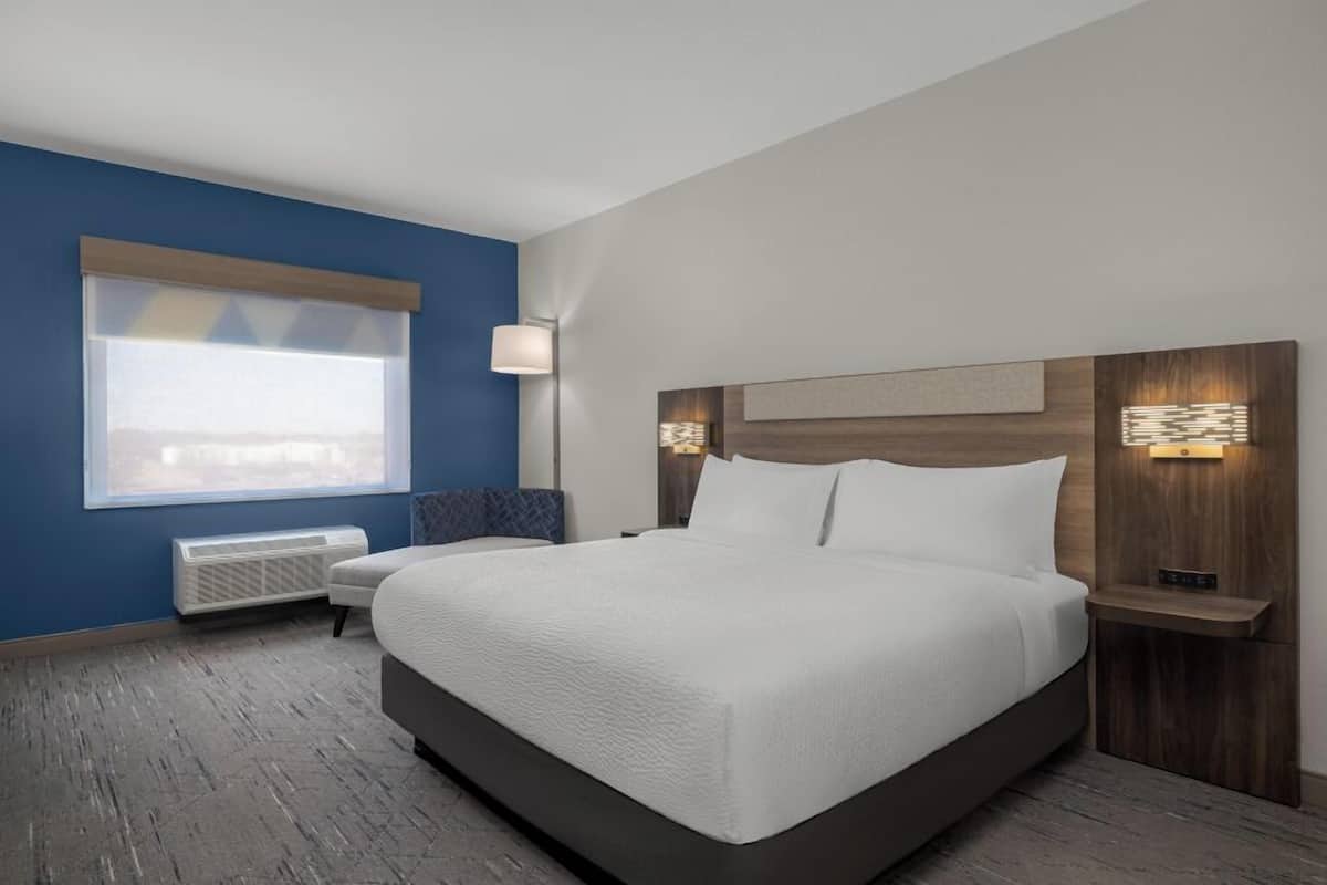 A hotel room with a large bed, a window with blinds, a bedside table with a lamp, and a wall-mounted light fixture.