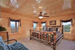 A wooden cabin bedroom with a rustic bed, a ceiling fan, and large windows offering a view of the outdoors. - Family-Friendly 12BR Lodge w/Games & Mtn. Views (Sevierville)