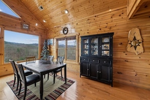 A wooden cabin interior with a dining table, chairs, a Christmas tree, and a cabinet with glass doors.