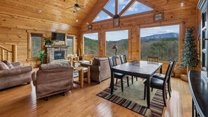 A log cabin interior with a fireplace, wooden furniture, a dining table, and large windows offering mountain views. - Cozy Cottage w/ Views, Hot Tub & Games (Sevierville)