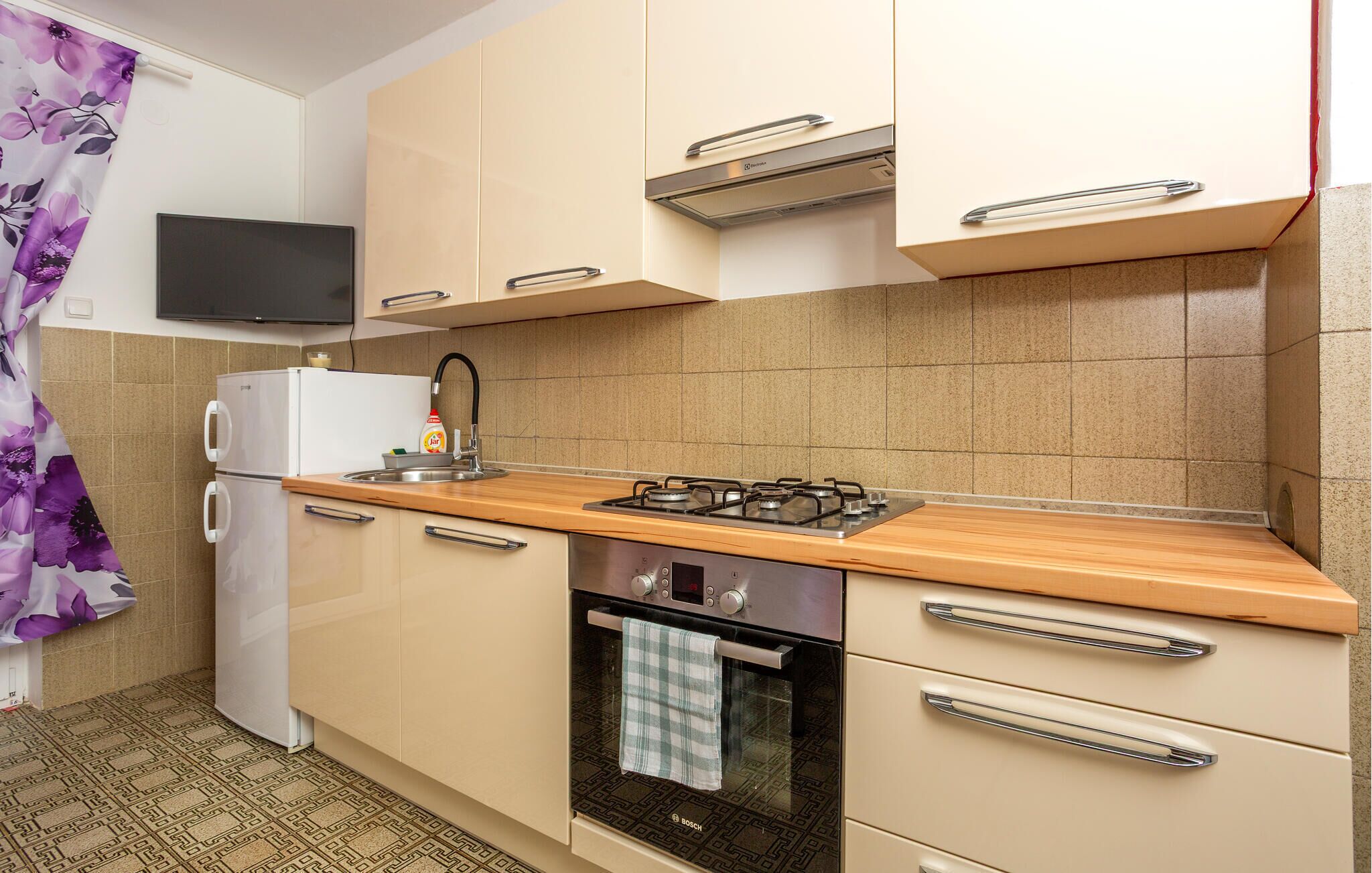 A compact kitchen with a refrigerator, stove, oven, and a flat-screen TV mounted above the counter.