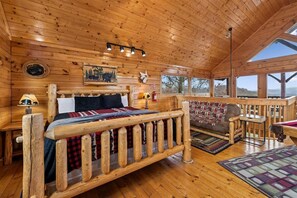A wooden cabin interior with a bed, a small table, a chair, and a view of the outdoors.