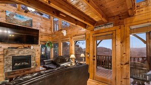 A log cabin interior with a fireplace, leather sofa, and a deck with a view of the mountains. - Luxury Cabin w/ Games, Views & Hot Tub (Sevierville)