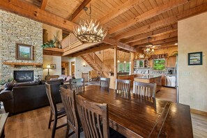 A spacious living area with a wooden dining table, comfortable seating, a stone fireplace, and a staircase leading to an upper level.