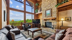 A spacious living room with leather sofas, a stone fireplace, a television, and a balcony with a view. - Grand Smoky Mountain Cabin w/ Views & Hot Tub (Sevierville)