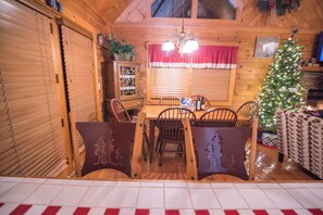 A cozy dining area with wooden chairs and a table, a Christmas tree, and a window with red curtains.