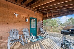 Una cabaña de madera con un porche, dos sillas Adirondack, una puerta verde y una parrilla para barbacoa.
