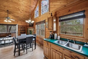 Interior de una cabaña de madera con comedor, cocina y amplias ventanas.