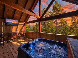 A wooden deck with a hot tub, seating area, and a view of trees and a sunset.