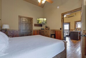 A bedroom with a large bed, wooden walls, a ceiling fan, and a view into the kitchen area.