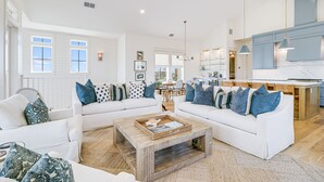 A spacious living room with white sofas, a coffee table, and a kitchen area in the background.