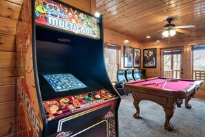 A pool table and a pinball machine in a room with wooden walls and ceiling, featuring a ceiling fan and framed pictures on the wall.