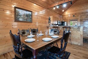 A wooden interior with a dining table set for six, a painting on the wall, and a kitchen area with cabinets and a microwave.