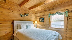 Quarto de cabana de madeira com uma cama, uma mesinha de cabeceira, uma luminária, uma janela com vista e um quadro na parede.
