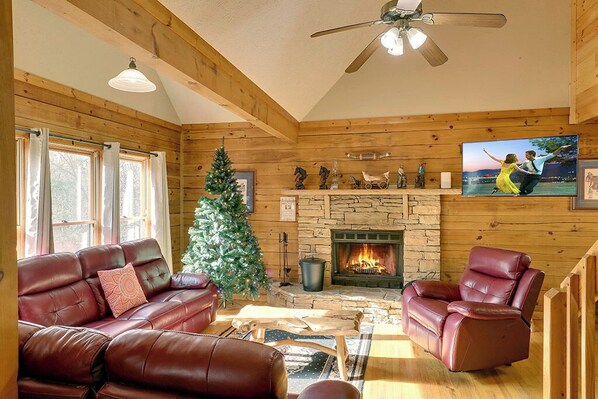 A cozy cabin interior with a fireplace, leather sofas, a Christmas tree, and a stone wall.