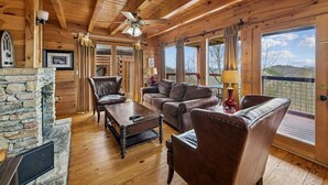 A log cabin interior with a stone fireplace, leather furniture, and large windows offering a view of the outdoors. - Charming Wooded Cabin w/ Game Area & Views (Sevierville)