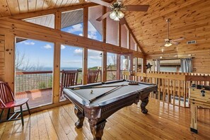 A wooden interior with a pool table, a ceiling fan, and large windows offering a view of the outdoors.