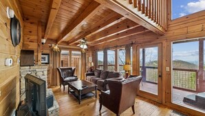 A wooden cabin interior with a fireplace, leather furniture, and large windows offering a view of the outdoors. - Charming Wooded Cabin w/ Game Area & Views (Sevierville)