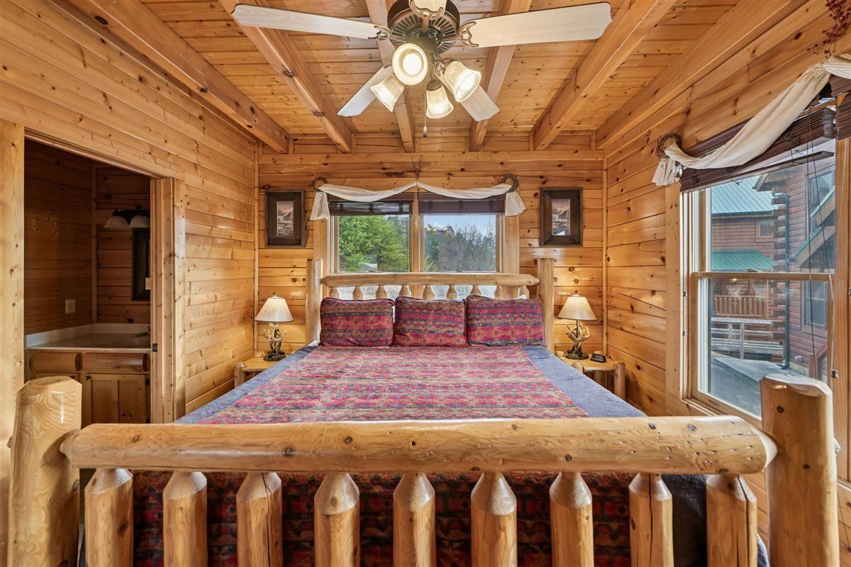 A wooden cabin bedroom with a bed, two bedside tables, a ceiling fan, and a window with curtains.
