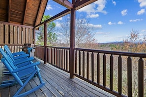 A wooden deck with blue rocking chairs, a wooden railing, and a view of trees and mountains.