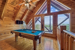 A wooden room with a pool table, large windows, and a ceiling fan.