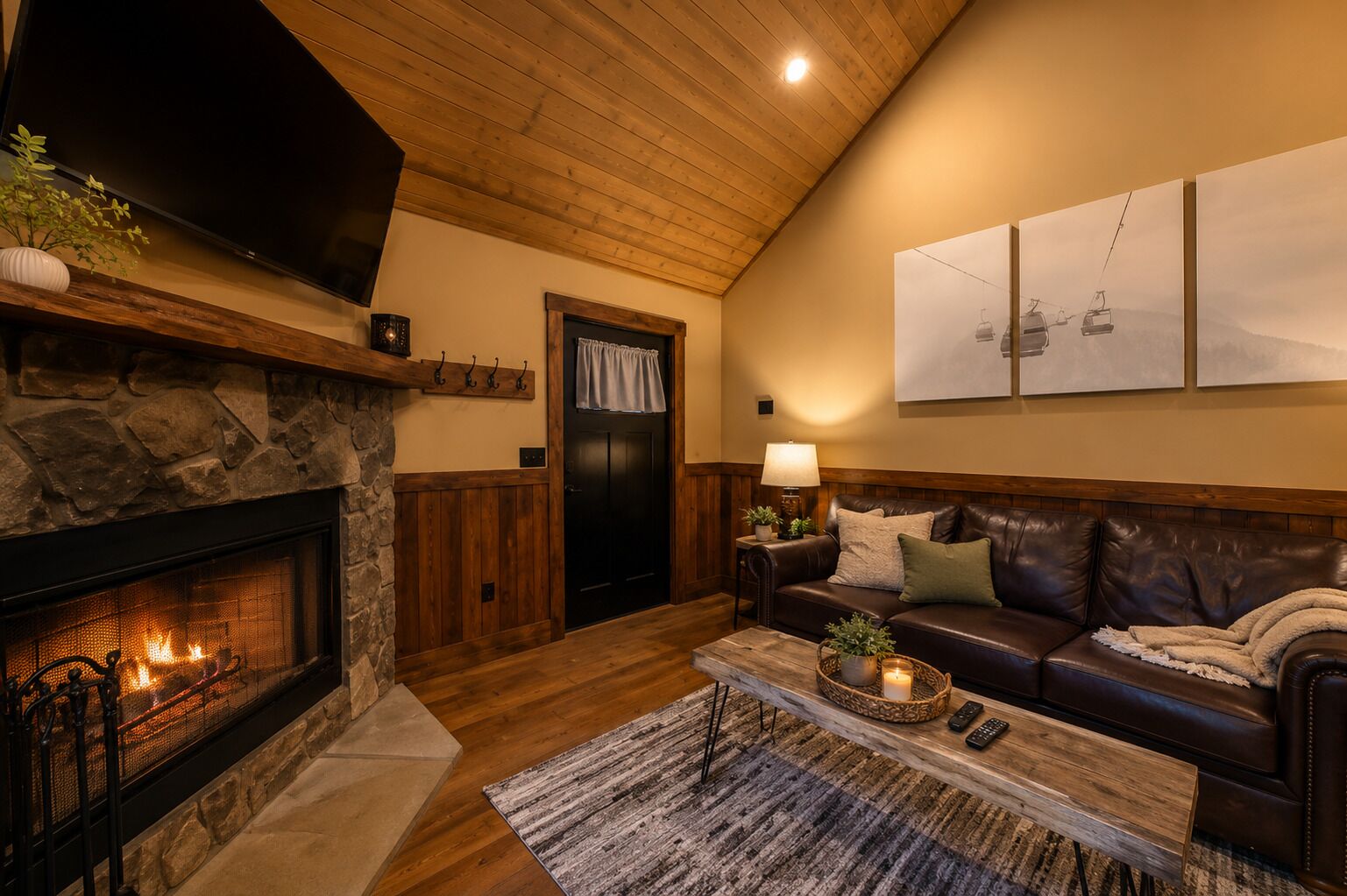 A cozy living room with a stone fireplace, a leather sofa, a coffee table with remote controls, and a wall-mounted TV.