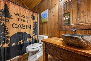 A cabin bathroom with a wooden interior, a welcome banner, a toilet, a stone sink, and a mirror. - Sevierville - Summit View - 1 Bdrms. (Sevierville)