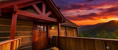 A wooden cabin with a wooden deck and a door, set against a backdrop of a sunset over a mountainous landscape.
