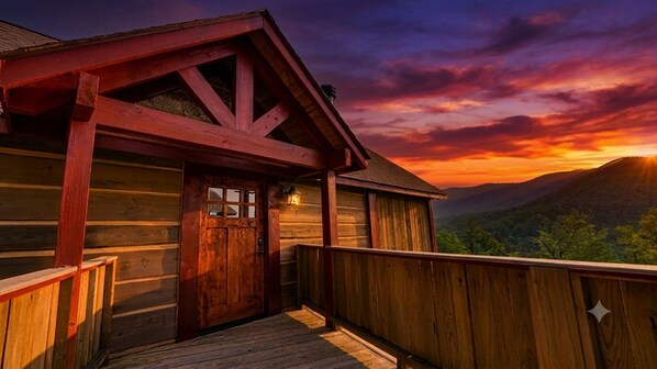 A wooden cabin with a wooden deck and a door, set against a backdrop of a sunset over a mountainous landscape.