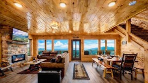 A cozy log cabin interior with a stone fireplace, a comfortable seating area, and a dining space with a scenic mountain view.